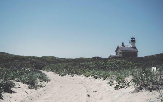 Scenic view of Block Island Lighthouse with sandy path and lush green dunes under blue sky.